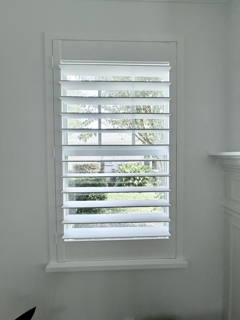 Interior view of a single window with custom white plantation shutters and 4.5” louvers, partially open to reveal a green garden outside.
