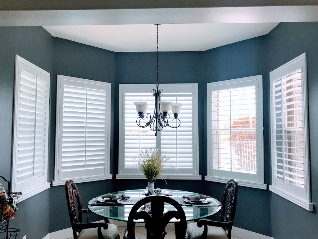 Dining room with a glass table surrounded by white plantation shutters on bay windows and deep navy blue walls.
