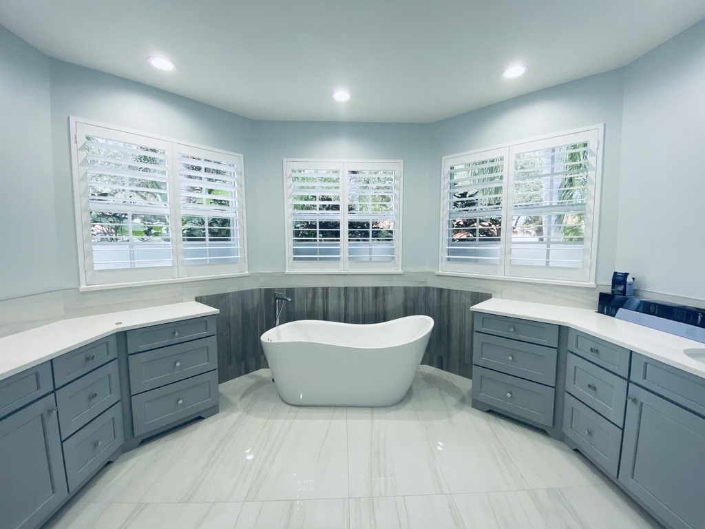 Elegant bathroom featuring a freestanding soaking tub surrounded by custom white plantation shutters on three corner windows, with gray cabinetry and marble flooring.